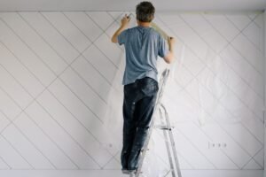 Full body back view of faceless male in shirt and dirty pants repainting wall while standing on ladder in bright empty room in daytime